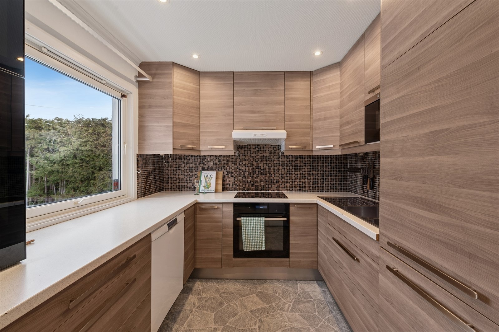 Kjøkkenet er utstyrt med IKEA-kjøkkeninnredning fra 2012, med glatte, laminerte fronter i imitert tre og takhøy løsning som gir godt med oppbevaringsplass. Benkeplatene er laminerte, og det er mosaikkfliser på vegg over arbeidsbenk som gir et flott preg. Galleribilde