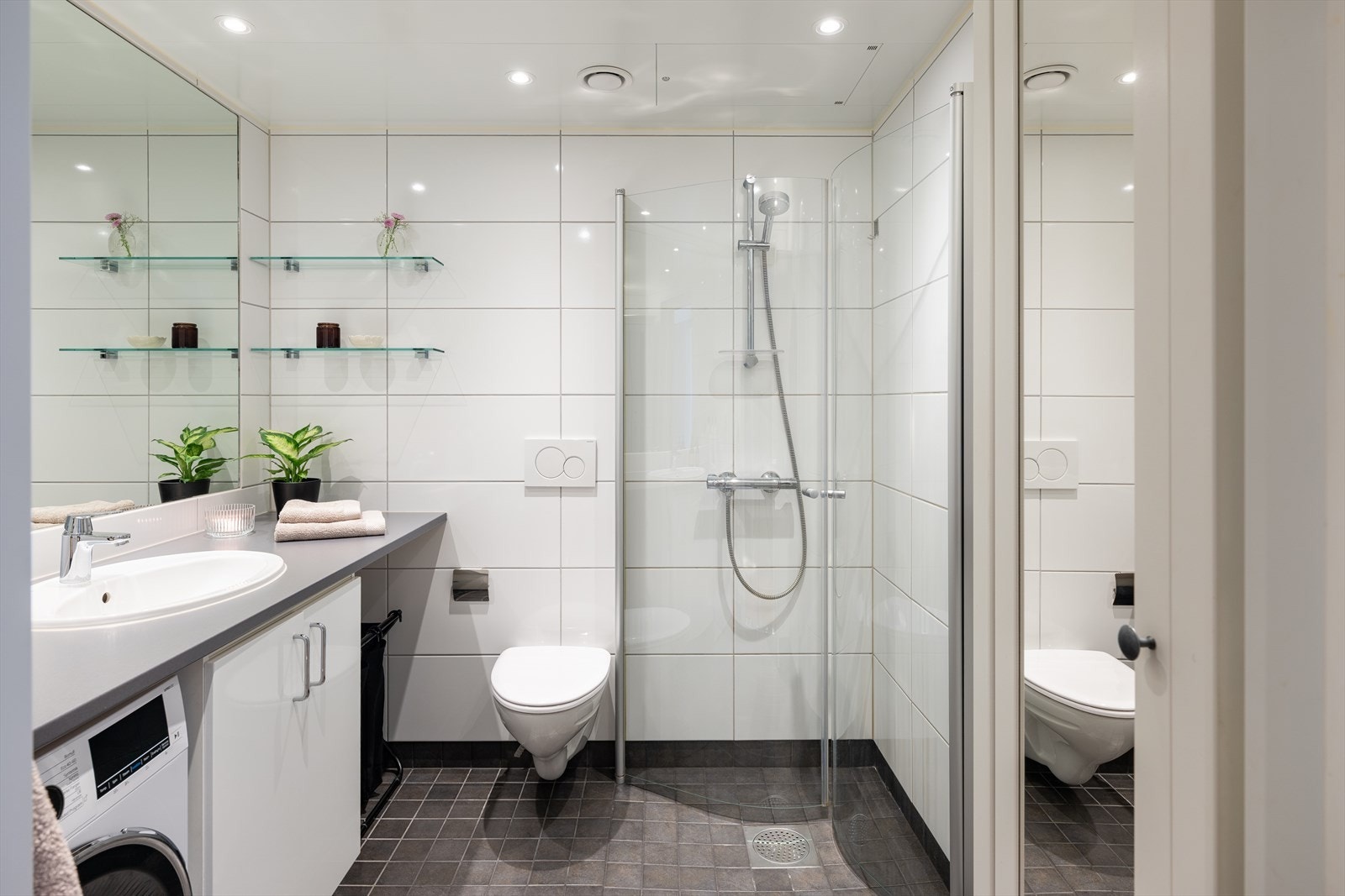 Badet er utstyrt med dusjhjørne med innfellbare glassdører, veggmontert toalett, servantskap med nedfelt servant og opplegg for vaskemaskin. Galleribilde