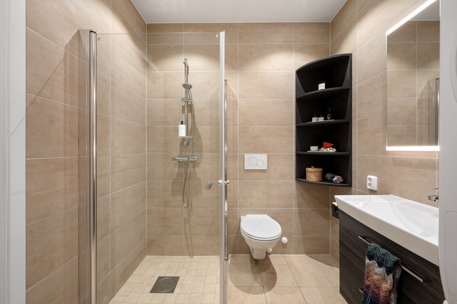 Badet er innredet med baderomsmøbel, heldekkende servant, speil med belysning, vegghengt toalett og dusjhjørne med innfellbare glassdører. Galleribilde