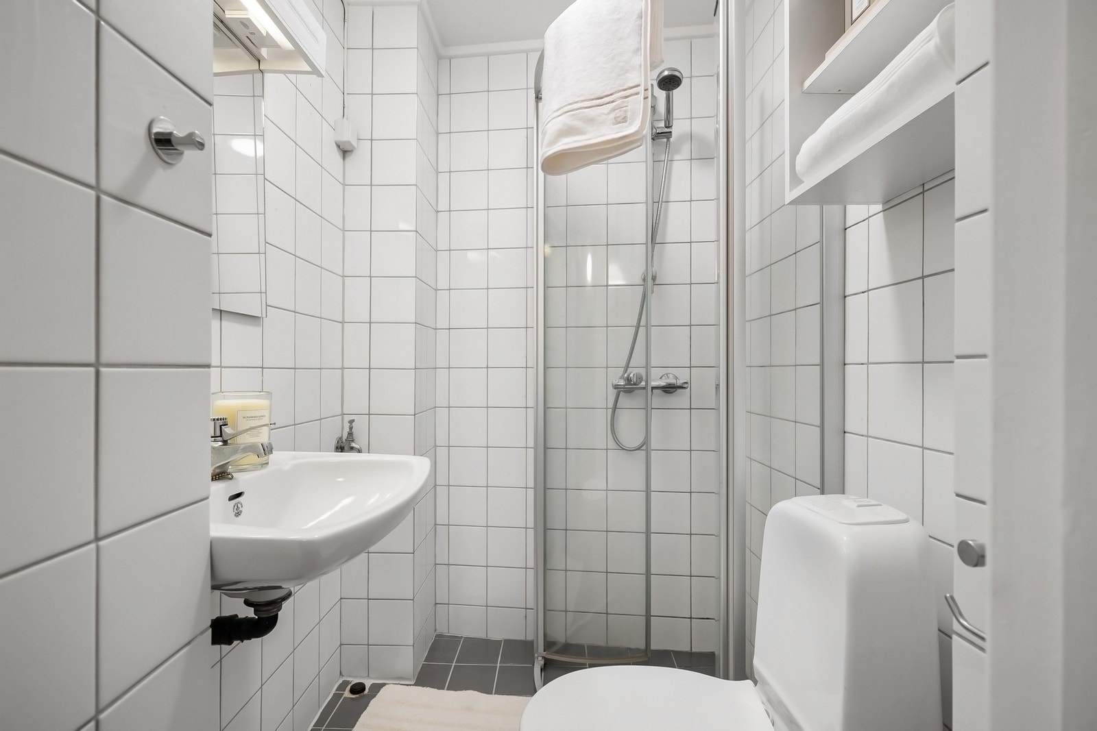 Badet er praktisk innredet med dusjhjørne med glassdører, gulvmontert toalett og servant med speil. Det er opplegg for vaskemaskin Galleribilde