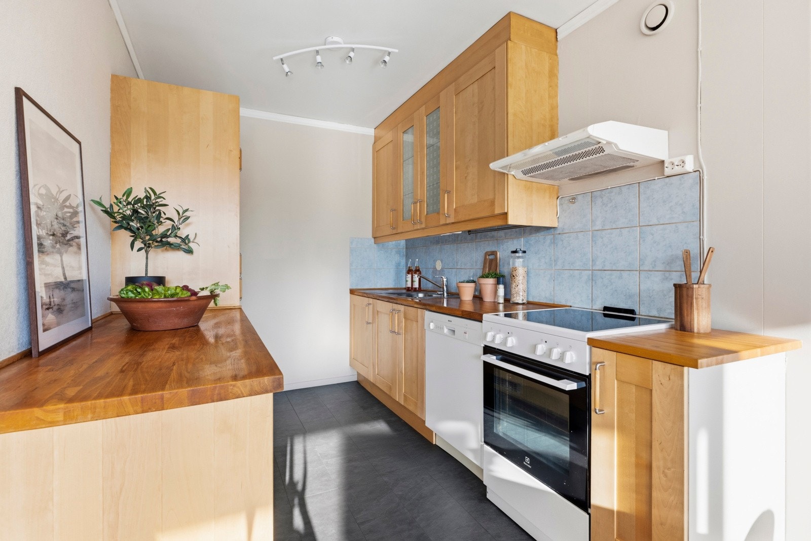 Innredningen har heltre/finerte fronter, eik/bøk benkeplater, oppvaskkum, ventilator, fliser over benk, belysning under overskap, oppvaskmaskin og komfyr med keramisk platetopp. Galleribilde