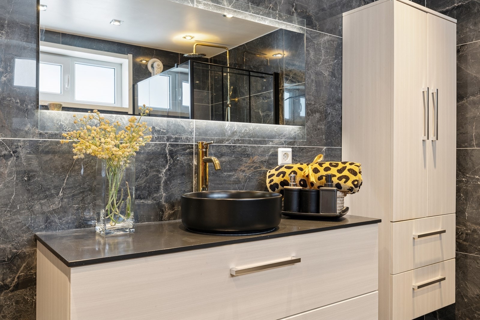 Badet er utstyrt med vegghengt toalett, dusjhjørne og en stor servantinnredning med speil, belysning og et praktisk veggmontert skap for praktisk oppbevaring. Galleribilde