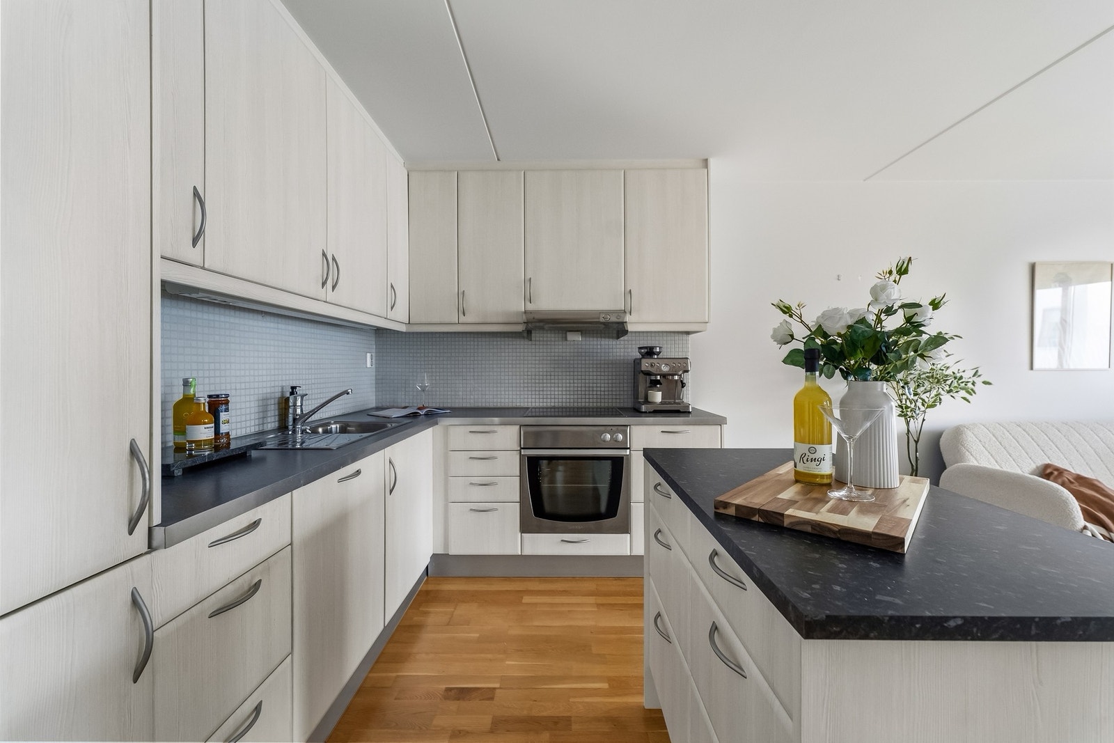 Kjøkkeninnredning med glatte fronter. Laminat benkeplate med nedfelt kum i rustfritt stål. Fliser på vegg over benkeskap. Kjøkkenøy i tilsvarende utførelse. Ventilator. Induksjonstopp, stekeovn, oppvaskmaskin og kjøl/frys er integrert i innredningen. Varm Galleribilde