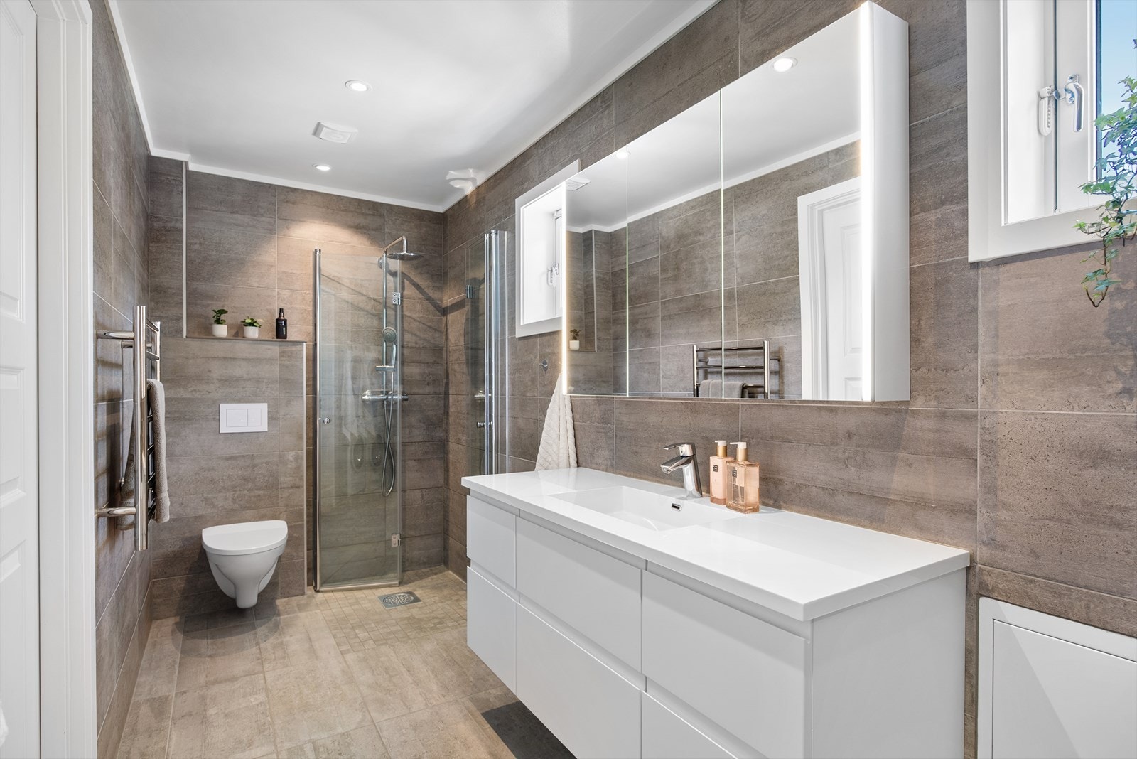 Videre er det dusjhjørne med innfellbare dusjvegger, vegghengt toalett og håndkletørker. Varmekabler i gulv og downlights i himling. Galleribilde