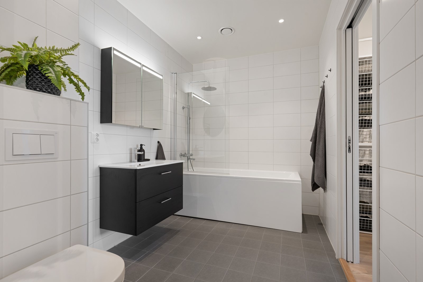 Lyst og innbydende bad med vegghengt toalett, helservant på skuffeseksjon, speil med skapplass og badekar Galleribilde