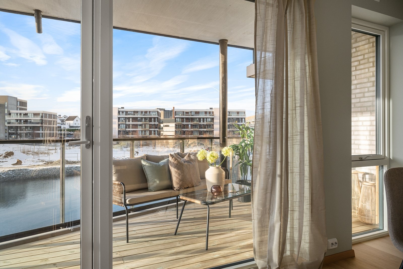 Fra stuen åpner dørene seg mot en solrik balkong med utsyn over vann og moderne bebyggelse. Galleribilde