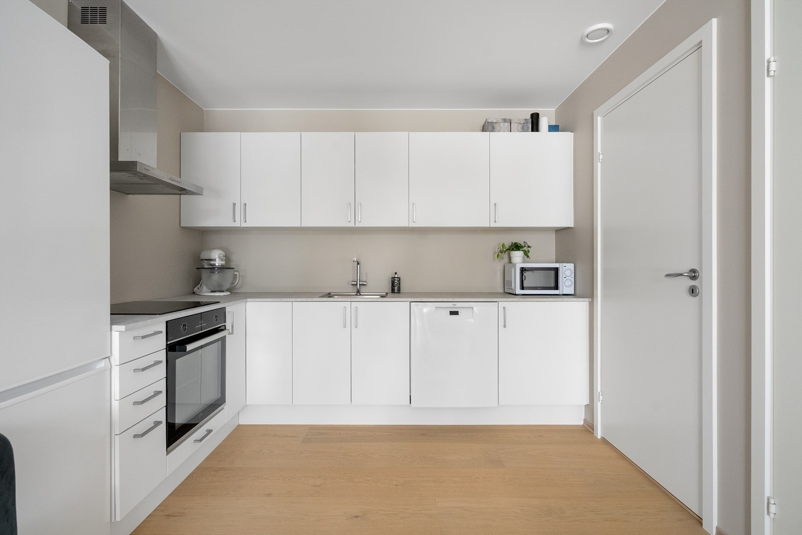 Kjøkken fra Sigdal med hvite, slette fronter og god skap- og benkplass. Kjøkkenet har integrert oppvaskmaskin, stekeovn og platetopp, samt frittstående kjøleskap. Galleribilde