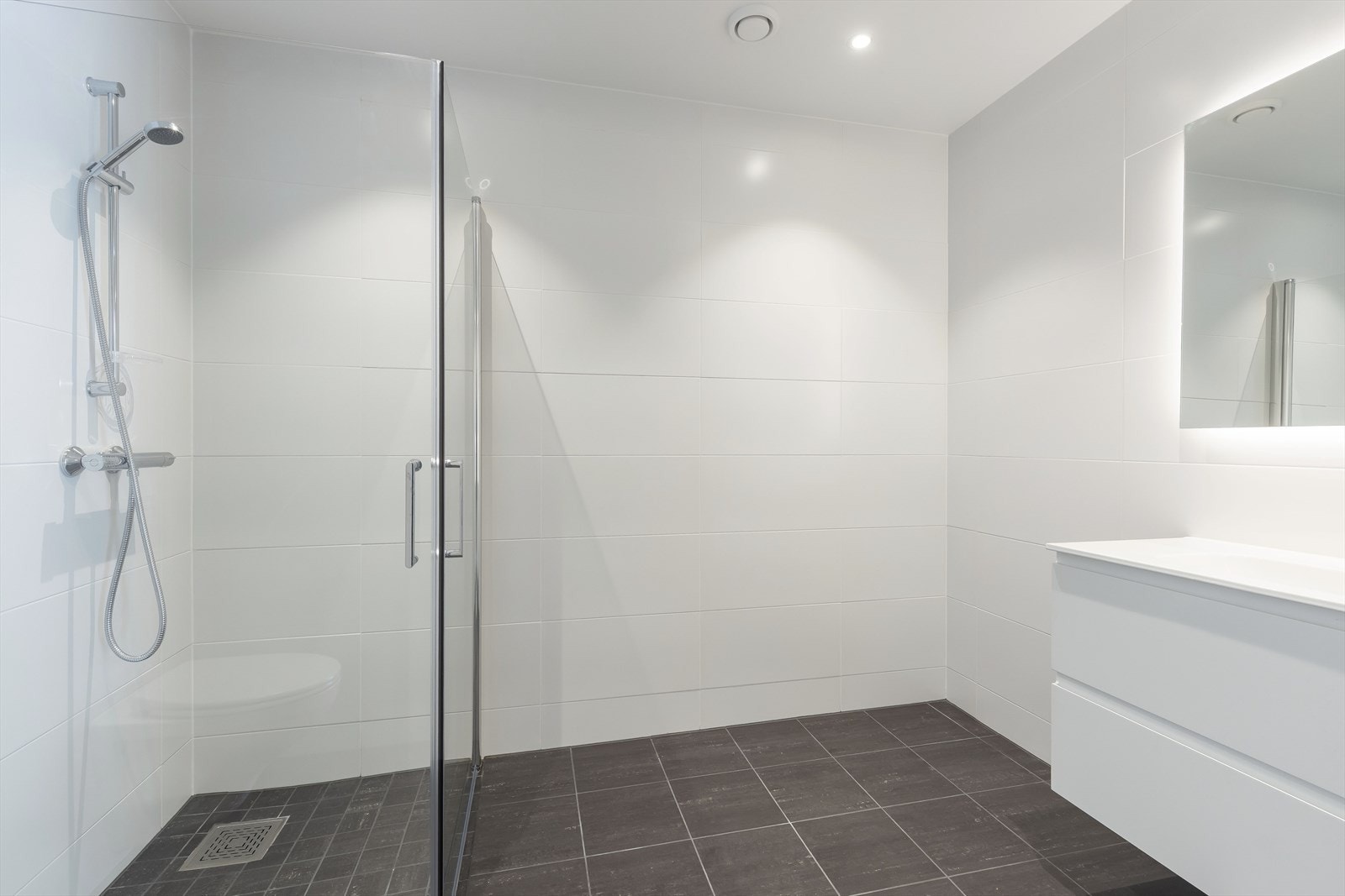 Badet er romslig og har flis på både vegger og gulv, badet har et speil med integrerte lys og en servantseksjon med skuffer Galleribilde
