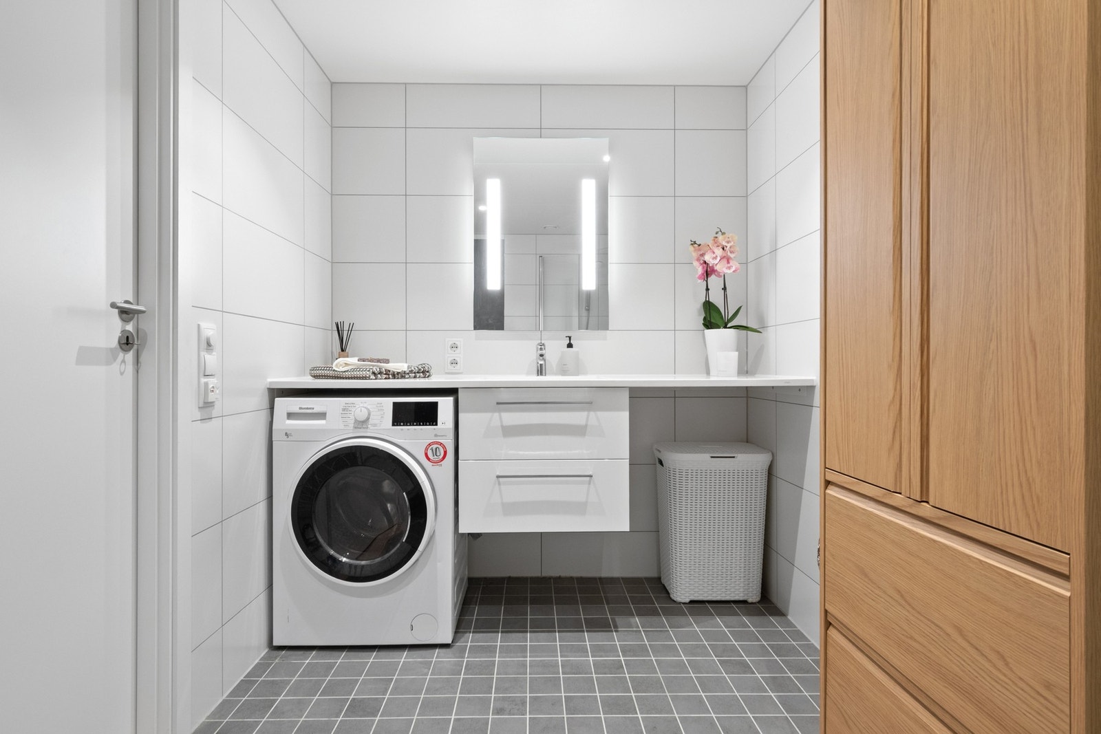 Romslig servantbenk med hvite skuffer og tilsvarende speilløsning. Under benken er det opplegg for vaskemaskin og plass til tørketrommel. Galleribilde