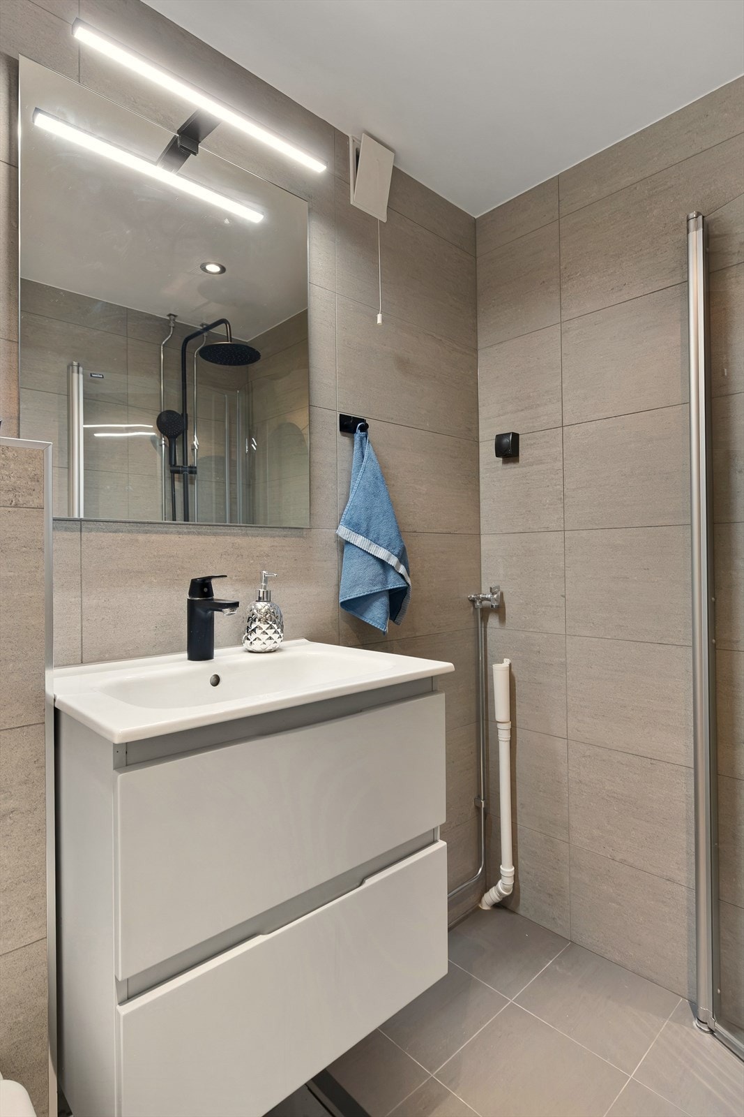 Flislagt bad med varmekabler i gulv og moderne innredning, dusjhjørne med innfellbare vegger og servant med skuffer. Galleribilde