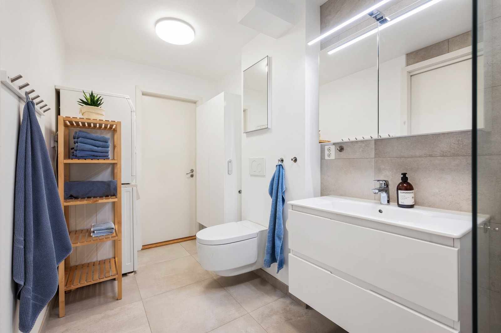 Tidløst flislagt bad med gulvvarme og opplegg for vaskemaskin. Videre er badet innredet med servantskap med speil med lys, vegghengt Geberit dusjttoalett og dusjhjørne. Galleribilde