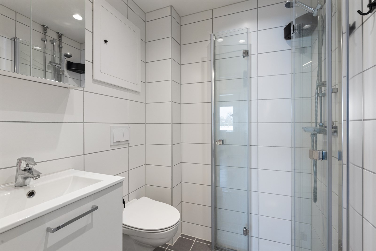 Flislagt bad/wc med varmekabler i gulv, servantskap, speil, vegghengt toalett og svingbare dusjdører. Galleribilde