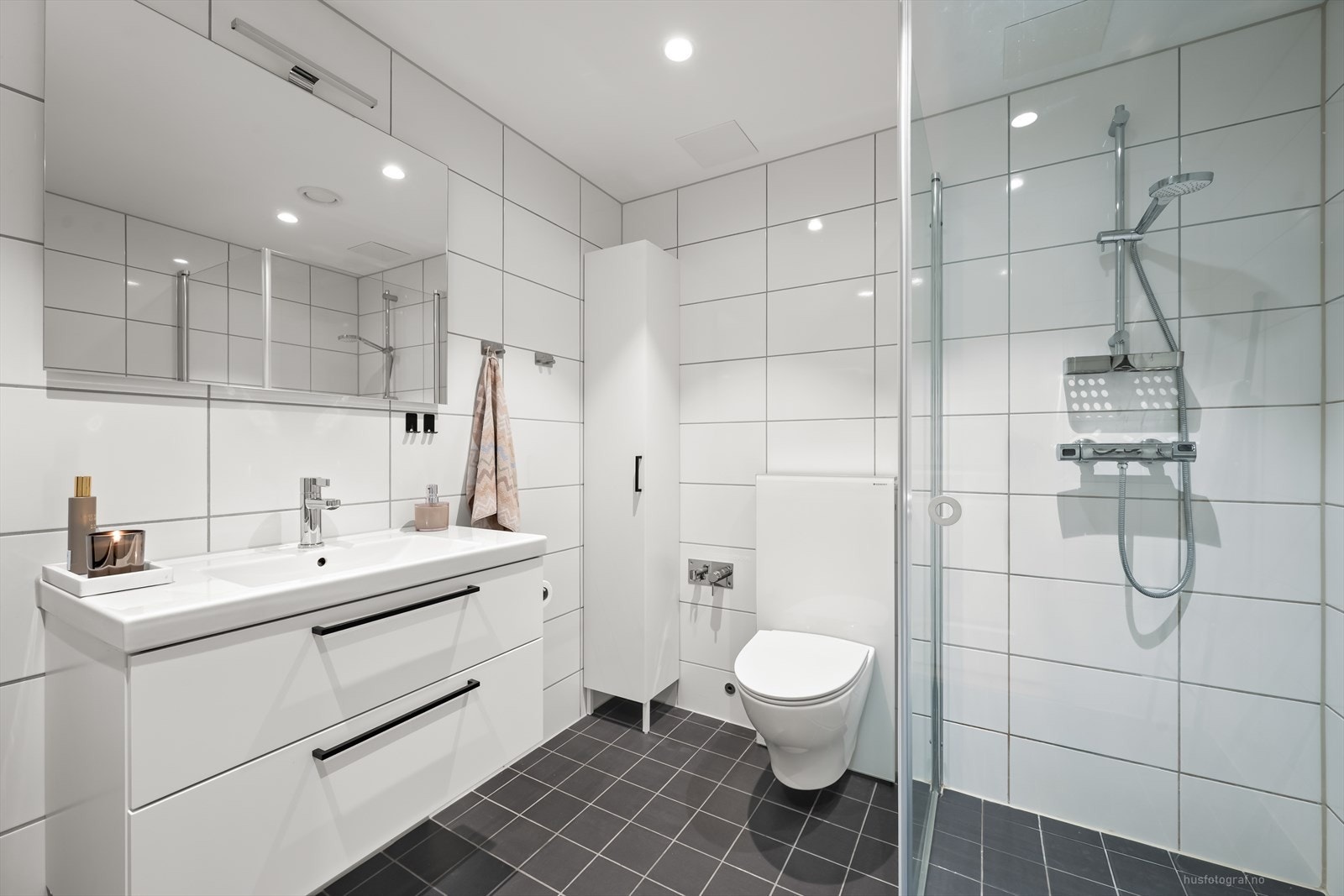 Leiligheten har et delikat baderom med gulvvarme og downlights i himling. Badet er innredet med et dusjhjørne med svingdører, veggmontert toalett, stilren innredning med heldekkende servant og speil med overliggende lys. Galleribilde