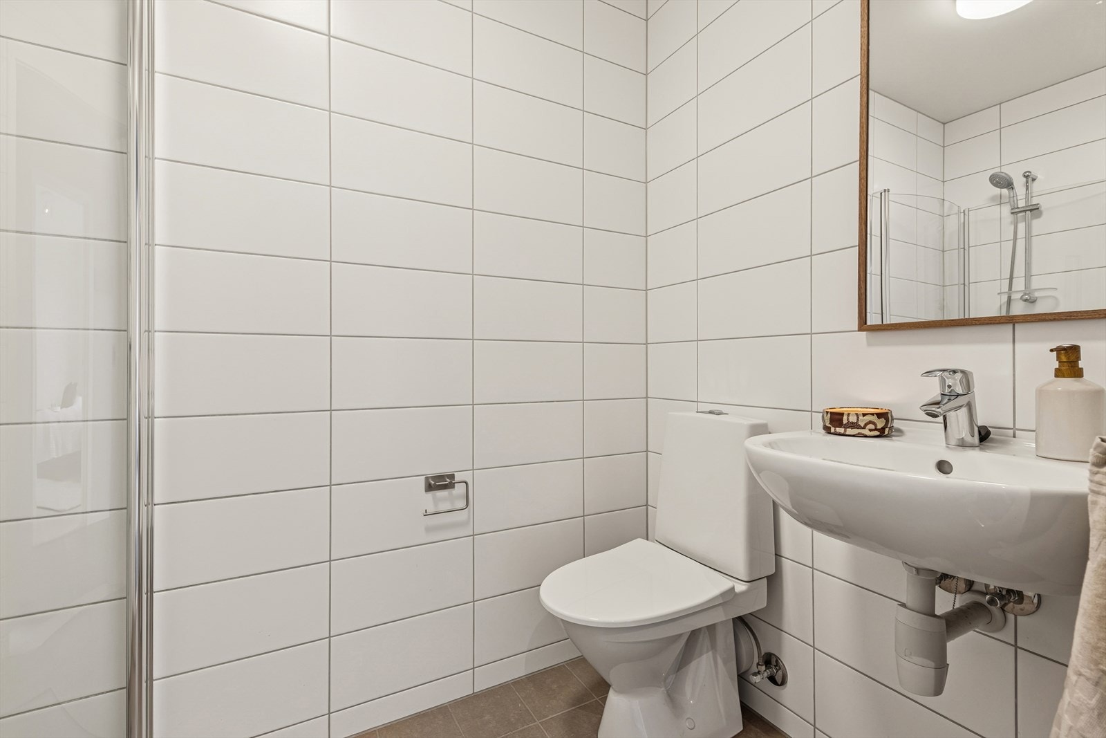 Baderommet i underetasjen er innredet i tilsvarende stil som badet i andre etasje med grå fliser på gulvet og hvite fliser på vegg. Også her har man vannbåren gulvvarme. Badet er ellers utstyrt med dusjhjørne, toalett og servant. Galleribilde