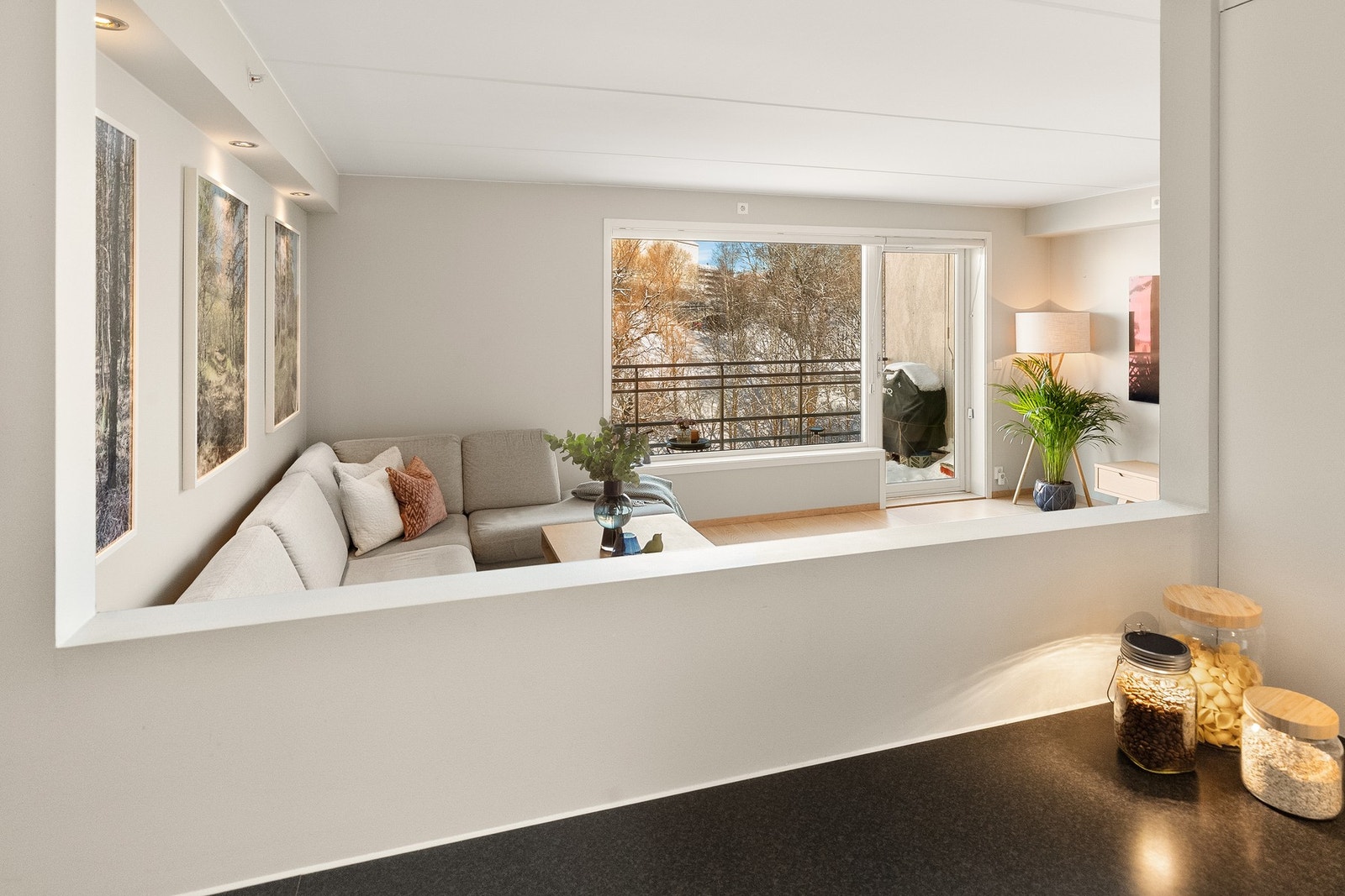 En veggåpning over benkehøyde gir visuell forbindelse til stuen og bidrar med lys og romfølelse, uten at kjøkkenet fremstår som helt åpent. Galleribilde