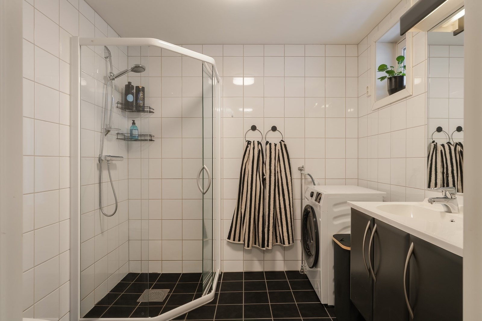 Flislagt bad med en ryddig og funksjonell utforming. Badet er utstyrt med dusjløsning med glassvegger, servant med skapplass og opplegg for vaskemaskin. Galleribilde