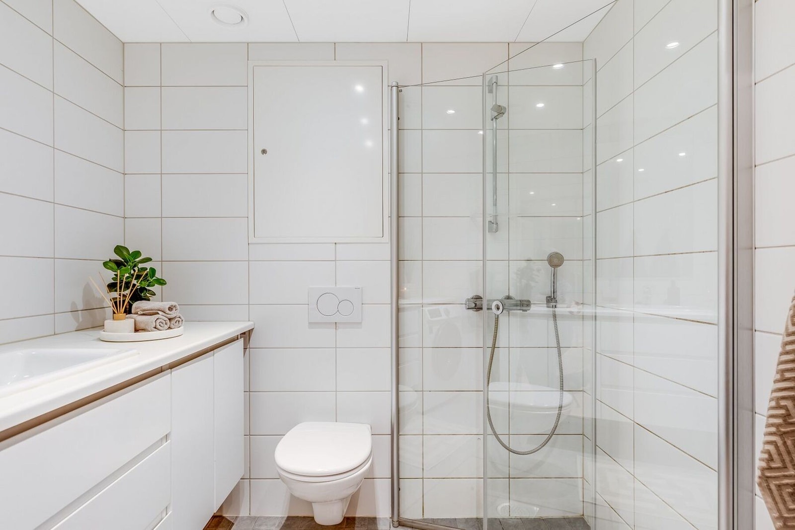Baderommet har egenskaper som dusjdører av glass, kran og avløp til vaskemaskin og veggmotert WC. Rørskapet er lokalisert over toalettet. Legg merke til servantplaten som strekker seg fra vegg til vegg og gir en praktisk løsning i hverdagen. Galleribilde