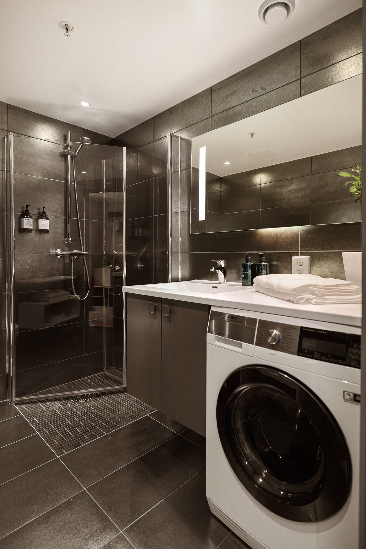 Moderne baderomsinnredning med heldekkende servant, stort speil med integrert belysning over. Praktisk med plass og opplegg for vaskemaskin under benkeplaten. Galleribilde