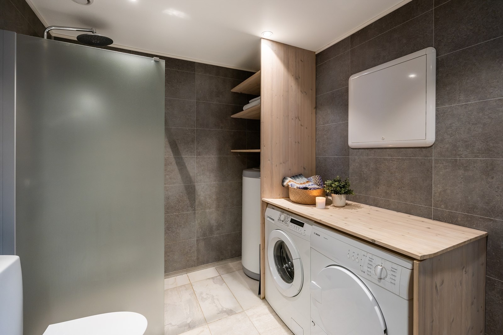 Badet er utstyrt med dusjvegg i glass, gulvmontert toalett, Innredning med servant og opplegg for vaskemaskin. Det er også en praktisk arbeidsbenk over vaskemaskin og tørketrommel. Galleribilde