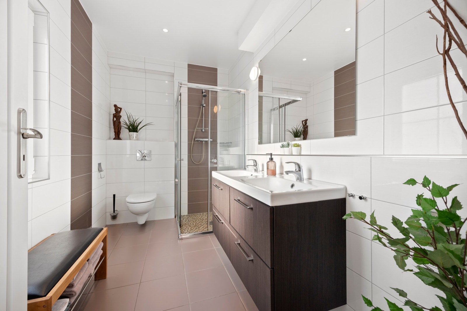 Pent bad med fliser på gulv og vegger. Innredet med vegghengt toalett, dusjhjørne med skyvedører og dobbel servant. Galleribilde