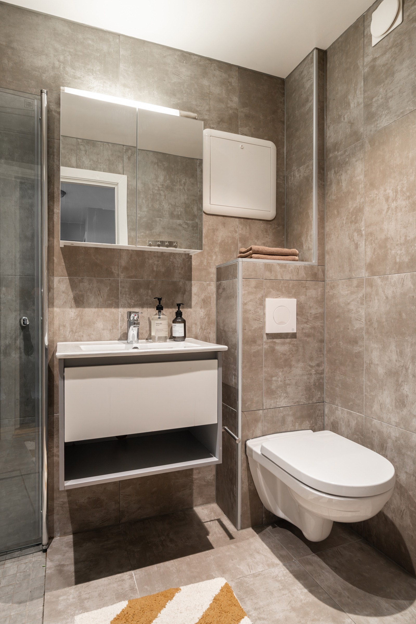 Stilren og lys baderomsinnredning med heldekkende servant, speilskap med belysning over. Baderommet er også utstyrt med dusjhjørne med innfellbare glassdører og vegghengt toalett. Galleribilde