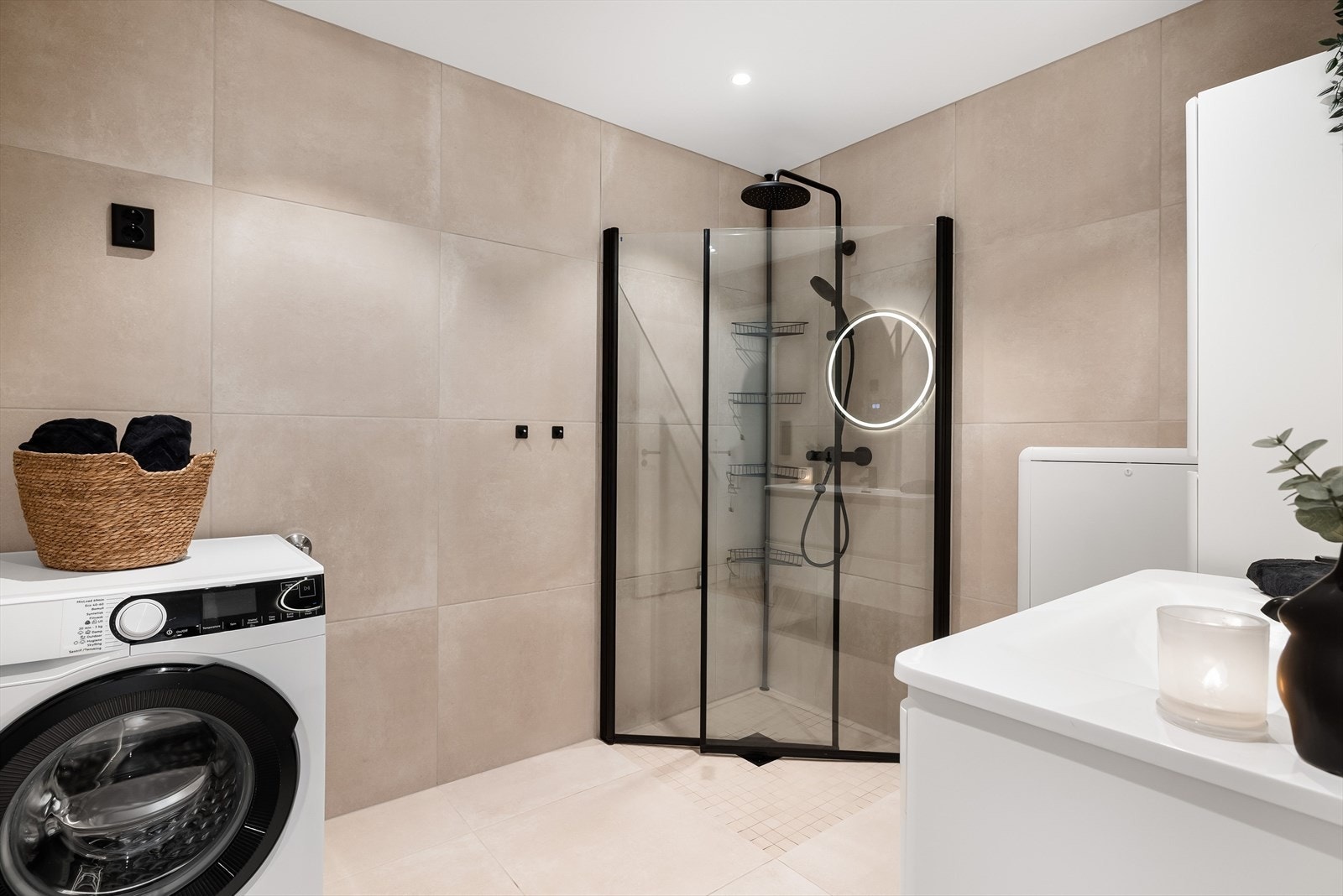 Badet har en praktisk dusjsone med mosaikkfliser på gulvet og innfellbare dusjdører. Det er montert vegghengt dusjbatteri med regnfall og en praktisk oppbevaringshylle i hjørnet. Galleribilde