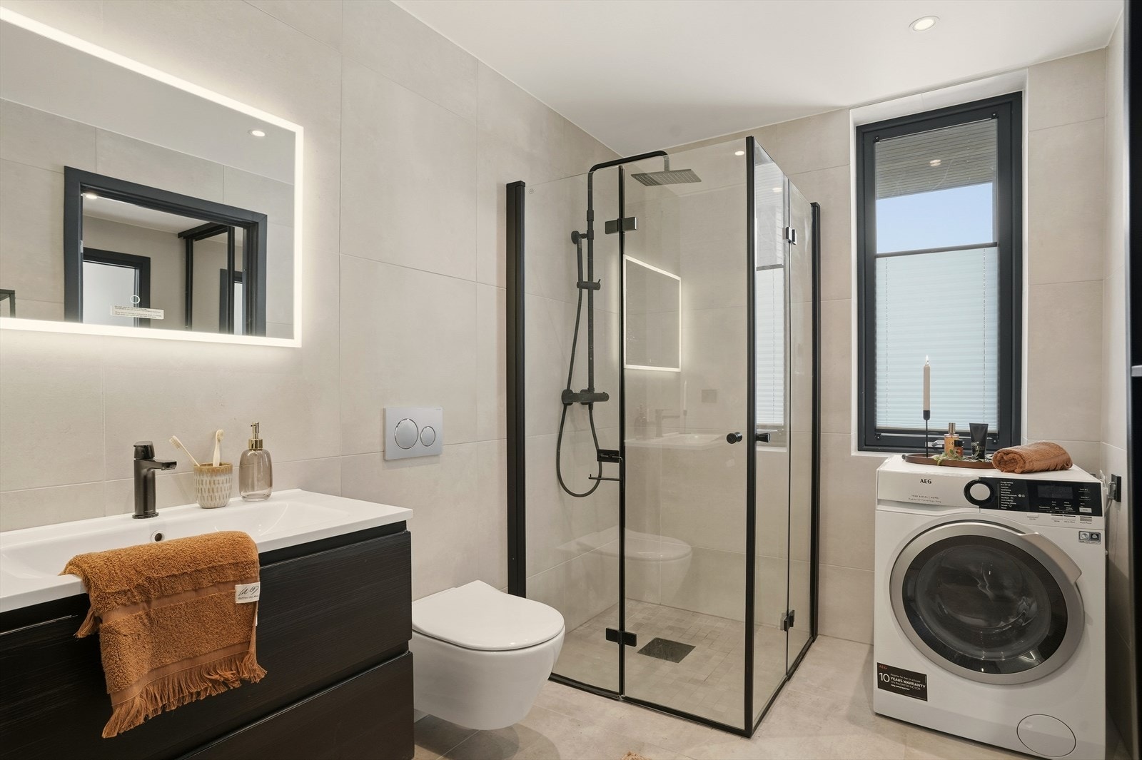 Praktisk bad/vaskerom med innredning og nedfelt servant, veggmontert toalett og dusjhjørne med glassvegger. Det er opplegg for vaskemaskin. Galleribilde