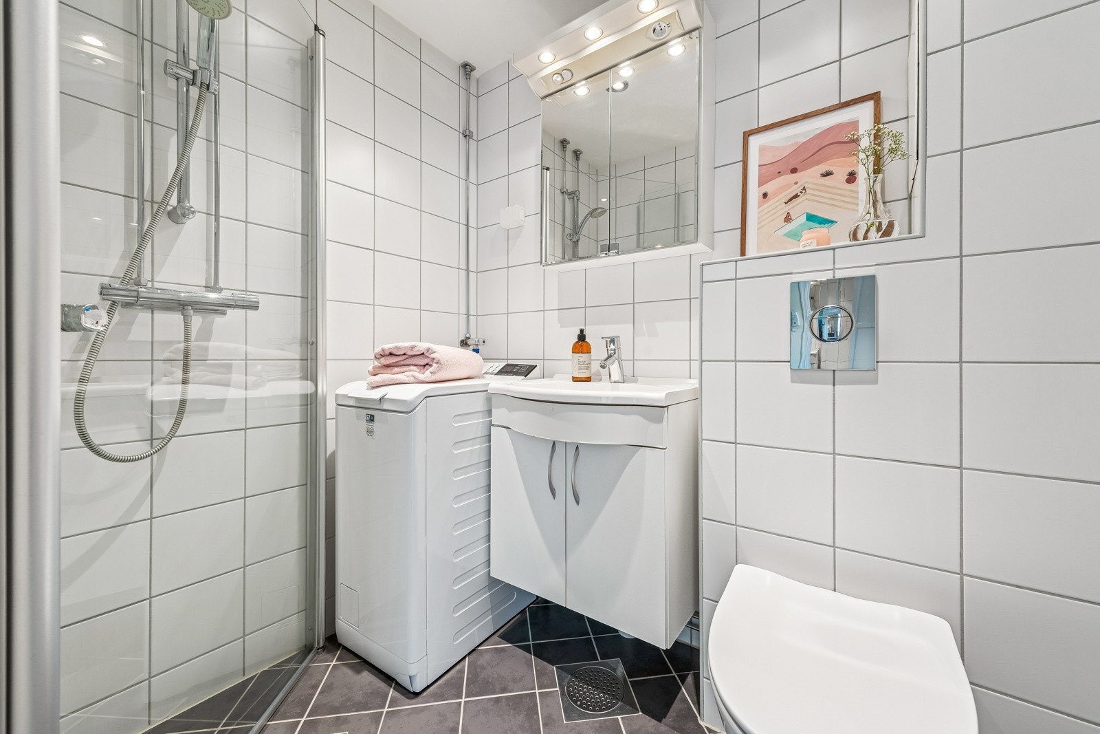 Badet er utstyrt med dusj med inn- utadslående dusjvegger, servant med underskap, overskap med speilfronter og lys over. Veggmontert klosett. Galleribilde