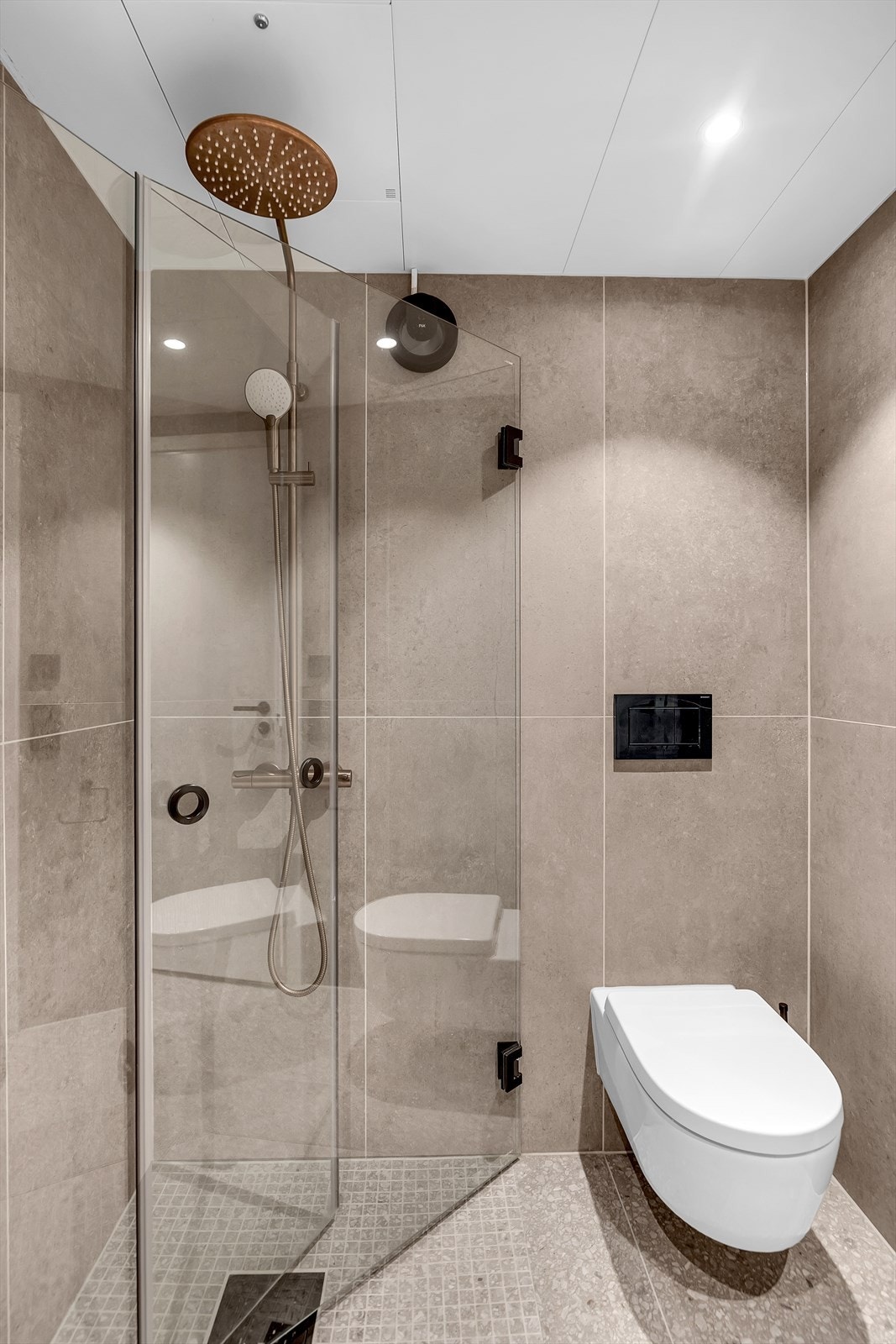 Dusjhjørne med mosaikkfliser på gulv, fossefalldusj og innfellbare glassdører, samt vegghengt toalett. Det er dimbare downligths i himling og varmekabler i gulv. Galleribilde