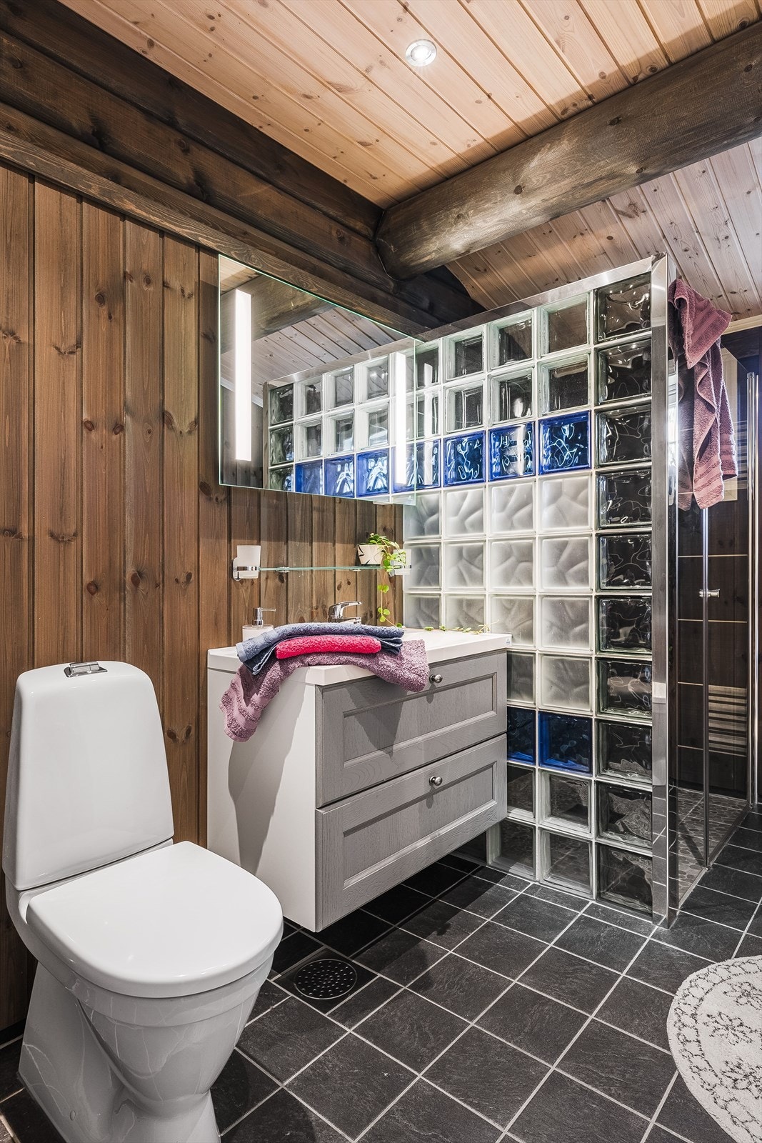 Pent baderom fra byggeåret 2015 med mørke fliser på gulv med gulvvarme, overflater med fliser og trepanel, downlights i himlingen og mekanisk avtrekksvifte. Galleribilde