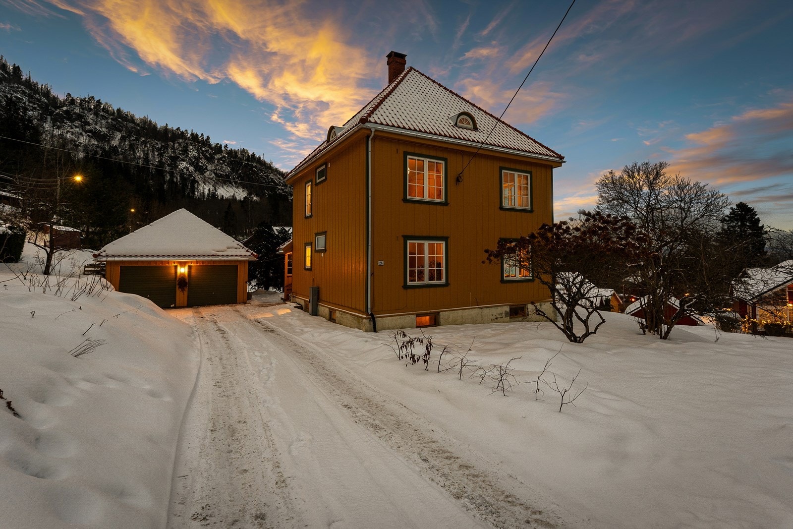 Parkering i frittstående garasje for to biler med elbil-lader fra Tesla. Det er stor sandkasse under snøen og frukttrær til venstre for innkjørselen. Galleribilde