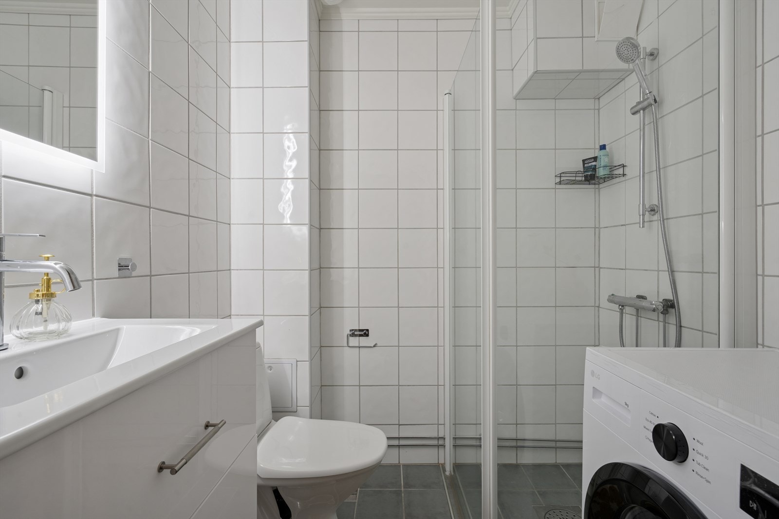 Badet har servant med ettgreps kran og skuffer under, speil med innebygget lys, gulvstående toalett og svingbare dusjdører i glass. Galleribilde