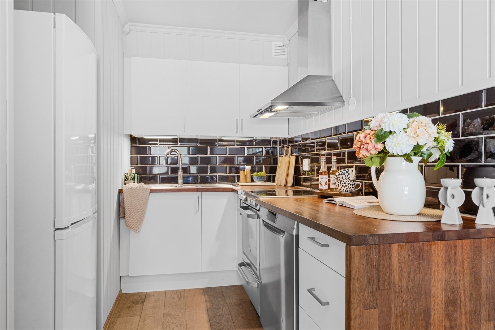 Kjøkkeninnredning med slette lyse fronter, flis over benk og det medfølger kjøleskap, oppvaskmaskin og komfyr Galleribilde