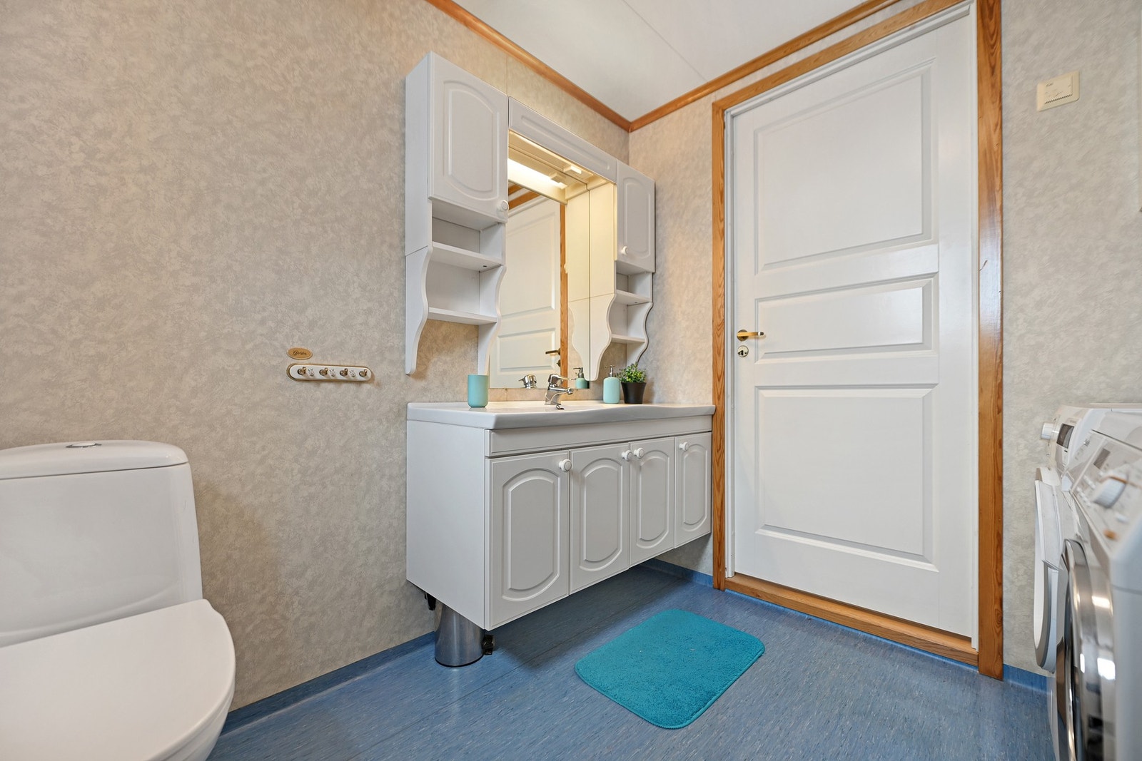 Badet har lyse vegger og blått gulv, med hvit innredning og speil over vasken. Galleribilde