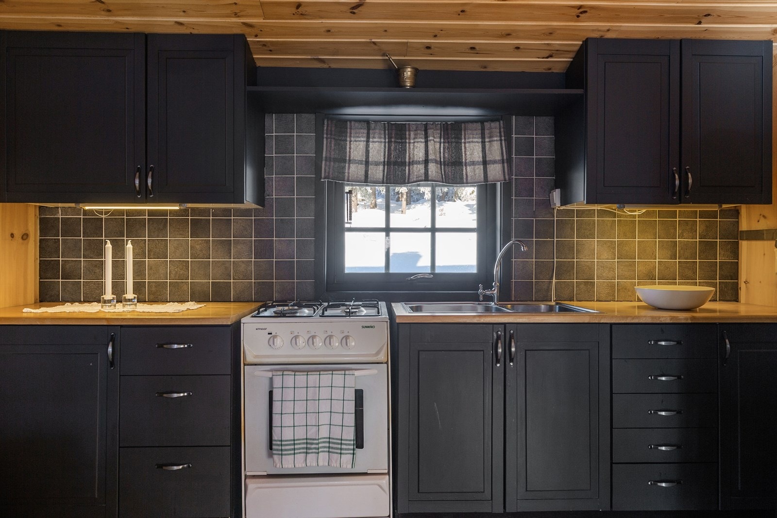 Det er montert fliser på vegg som gir en fin kontrast til de mørke kjøkkenskapene. Her finner du nedfelt oppvaskkum, frittstående gasskomfyr og gasskjøle-/fryseskap. Galleribilde