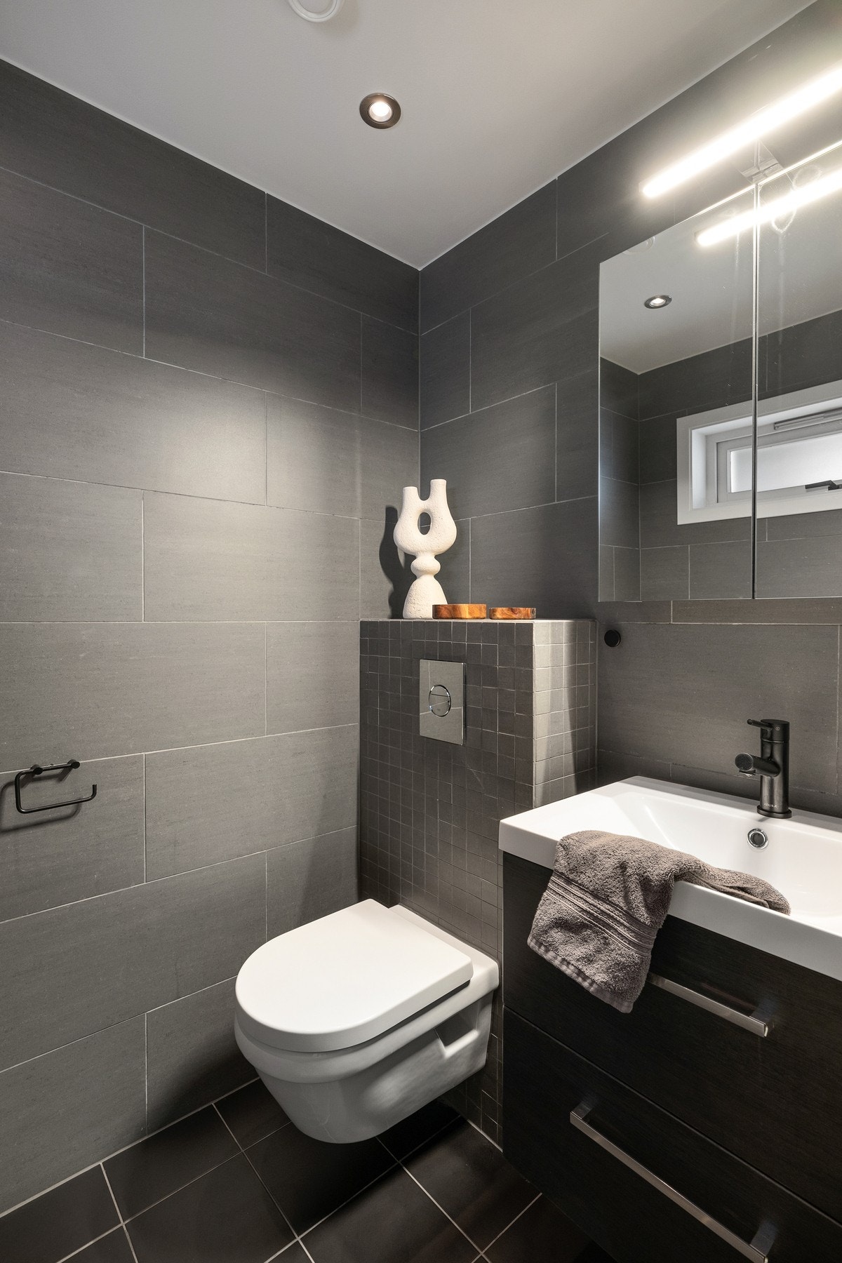I tilknytning til inngangspartiet ligger et gjestetoalett med mørke fliser og vegghengt toalett, en praktisk løsning for både familie og besøkende. Galleribilde