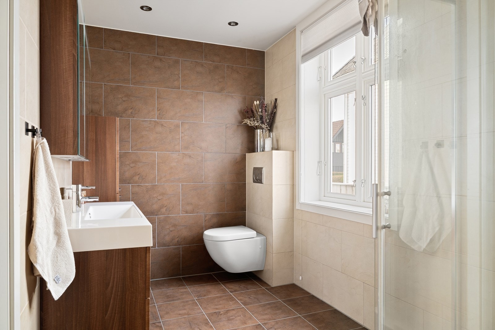 Badet fremstår elegant med fliser på både gulv og vegger. Her finner du dusjhjørne med regndusj, vegghengt toalett, servant med underskap og speil med integrert belysning - alt tilrettelagt for en moderne og komfortabel hverdag. Galleribilde