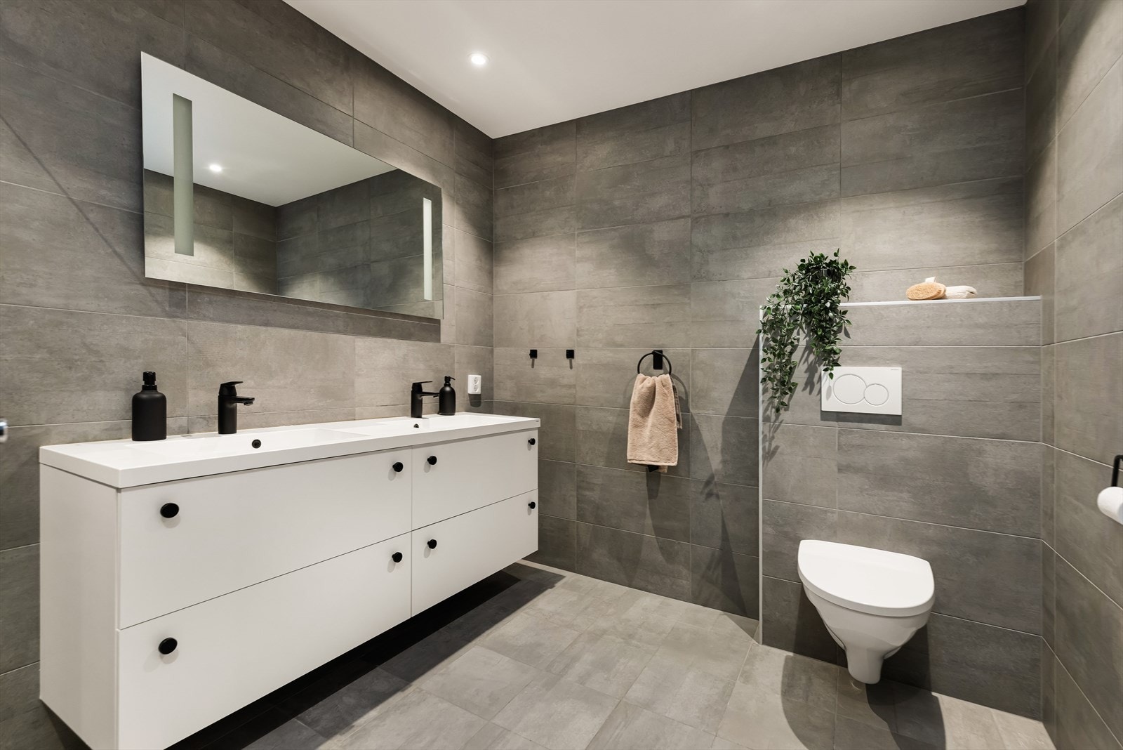 Stilrent bad med benkeskap med nedfelt servant og speil med integrert belysning. Badet er utstyrt med vegghengt toalett og dusjhjørne. Galleribilde