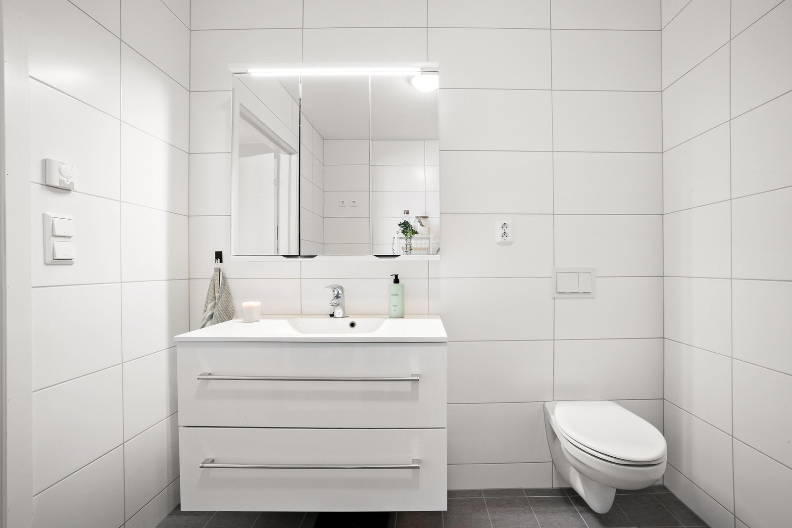 Badet har moderne hvite fliser, vegghengt toalett og servant med oppbevaringsskuffer. Galleribilde