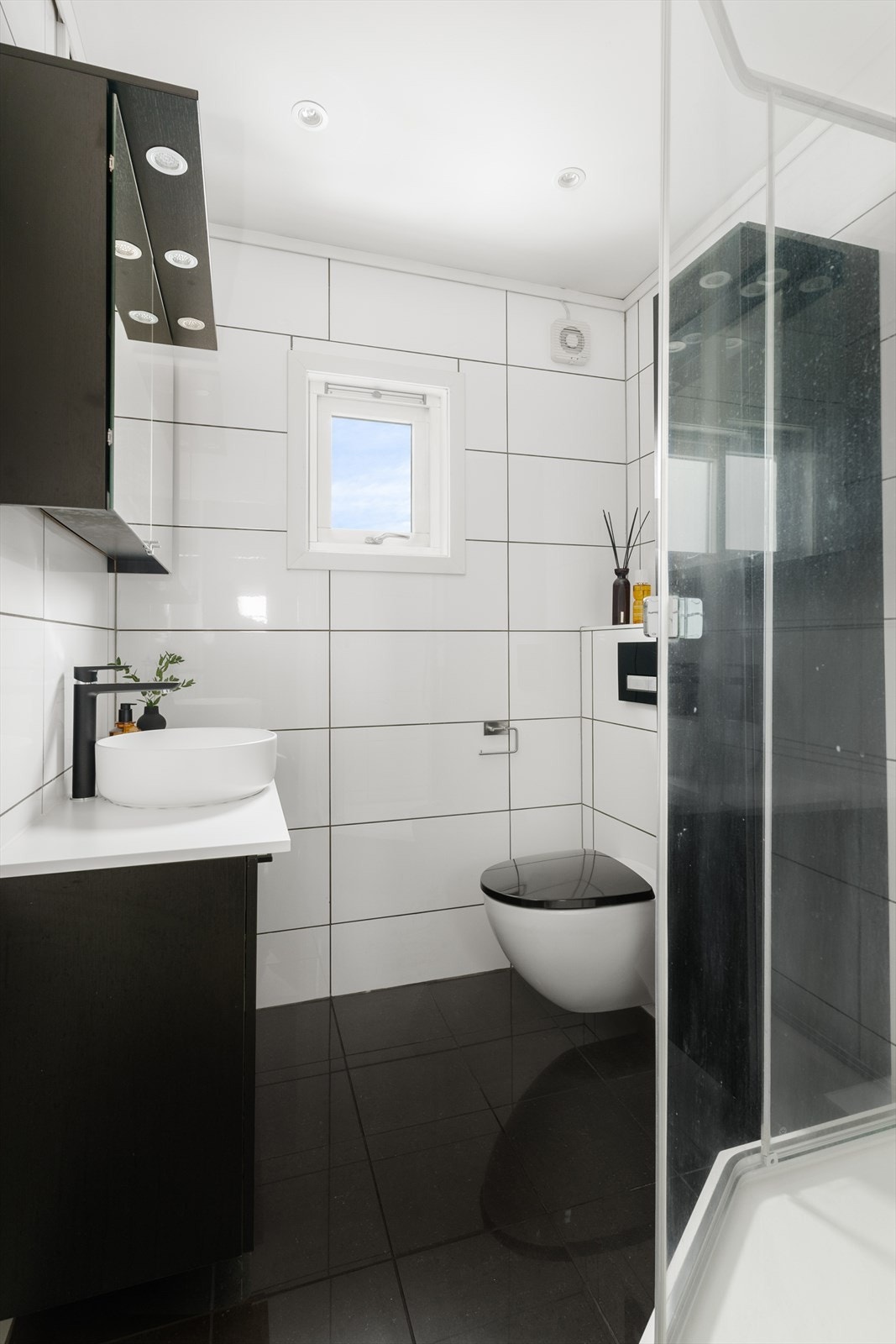 Badet er innredet med servantskap og høyskap i mørk utførelse, samt speilskap med overlys. Servant, benkeplate, blandebatteri og vannlås er nytt i 2026. Badet har også dusjkabinett og veggmontert toalett. Galleribilde