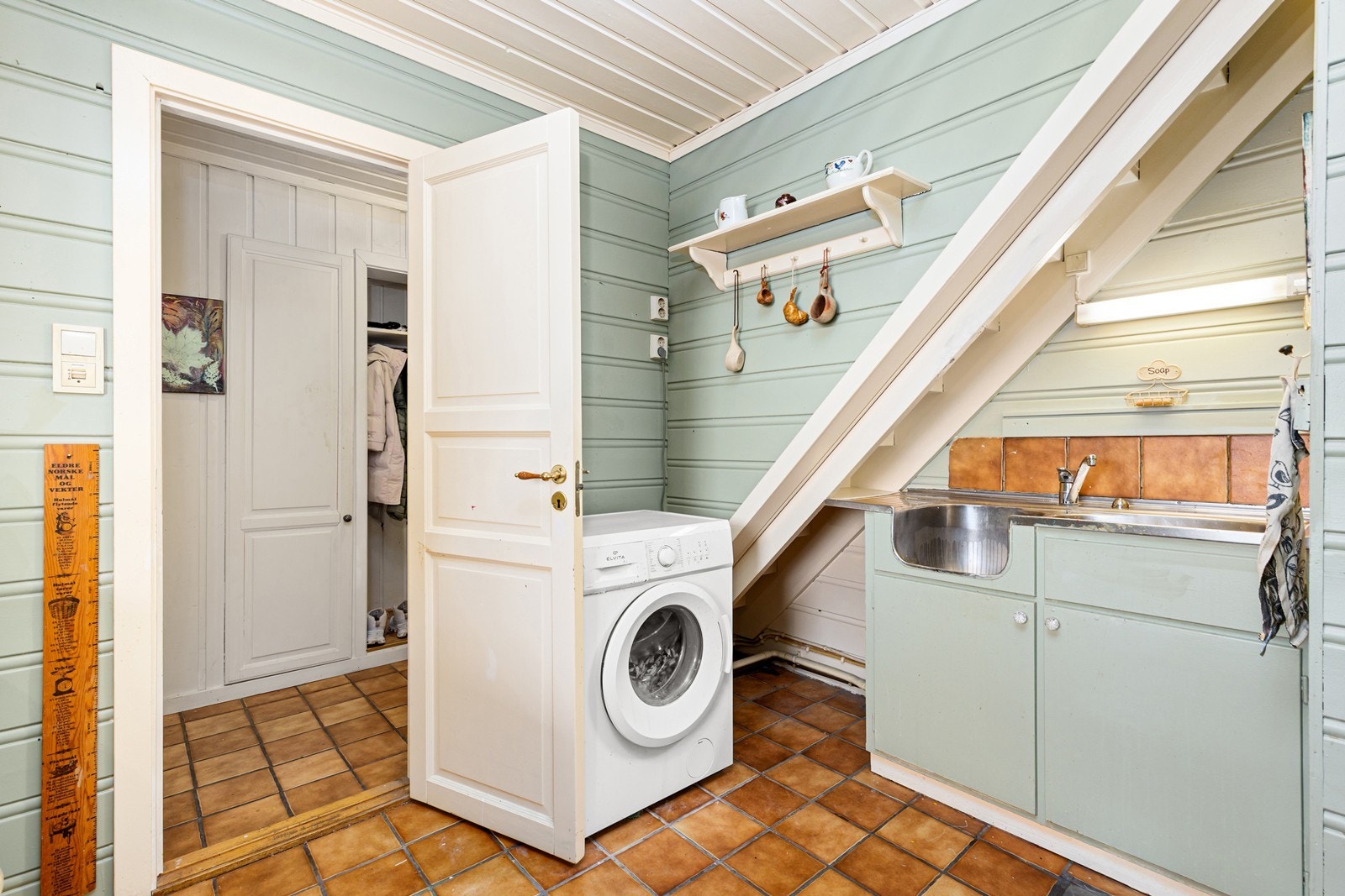 Enkelt, eldre vaskerom med flislagt gulv og malt panel på veggene. Det er sluk av støpejern i gulvet, eldre skapinnredning og opplegg for vaskemaskin. Galleribilde