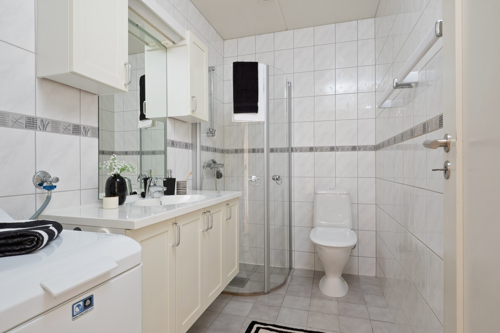 Eldre bad med fliser på gulv og elektrisk gulvvarme. Rommet er innredet med servantinnredning, dusjdører, wc og opplegg for vaskemaskin. Galleribilde