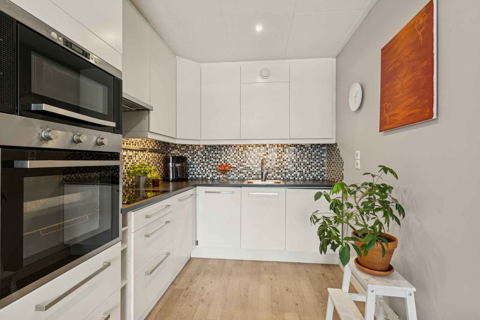 Kjøkkeninnredning med under- og overskap med slette fronter, laminatbenkeplate med nedfelt kum i rustfritt stål samt integrerte hvitevarer bestående av platetopp, stekeovn, mikrobølgeovn, oppvaskmaskin og kjøl/frys. Galleribilde