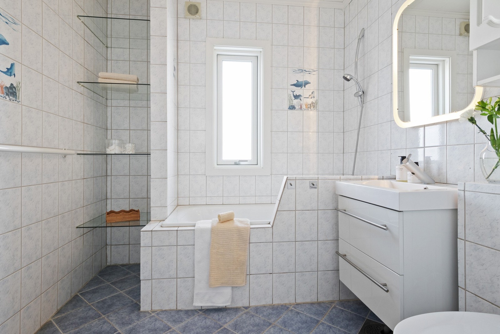 Badet har badekar har flislagte overflater, veggnisje med glasshyller og hvit baderomsinnredning med heldekkende servant. Galleribilde