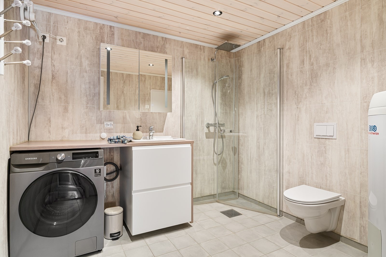 Bad/vaskerom med med fliser på gulv med varmekabler under. Praktisk løsning med vask i seksjon, benkeplate med vaskemaskin under. Dusj med fleksible dusjdører. Veggheng wc. Galleribilde