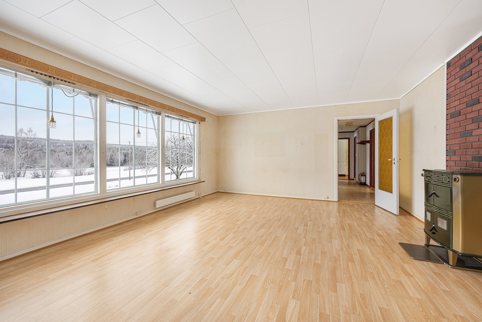 Rommet har lys tapet på veggene og belegg i parkettimitasjon på gulvet, og i tillegg til panelovner har stuen en eldre kombiovn. Galleribilde