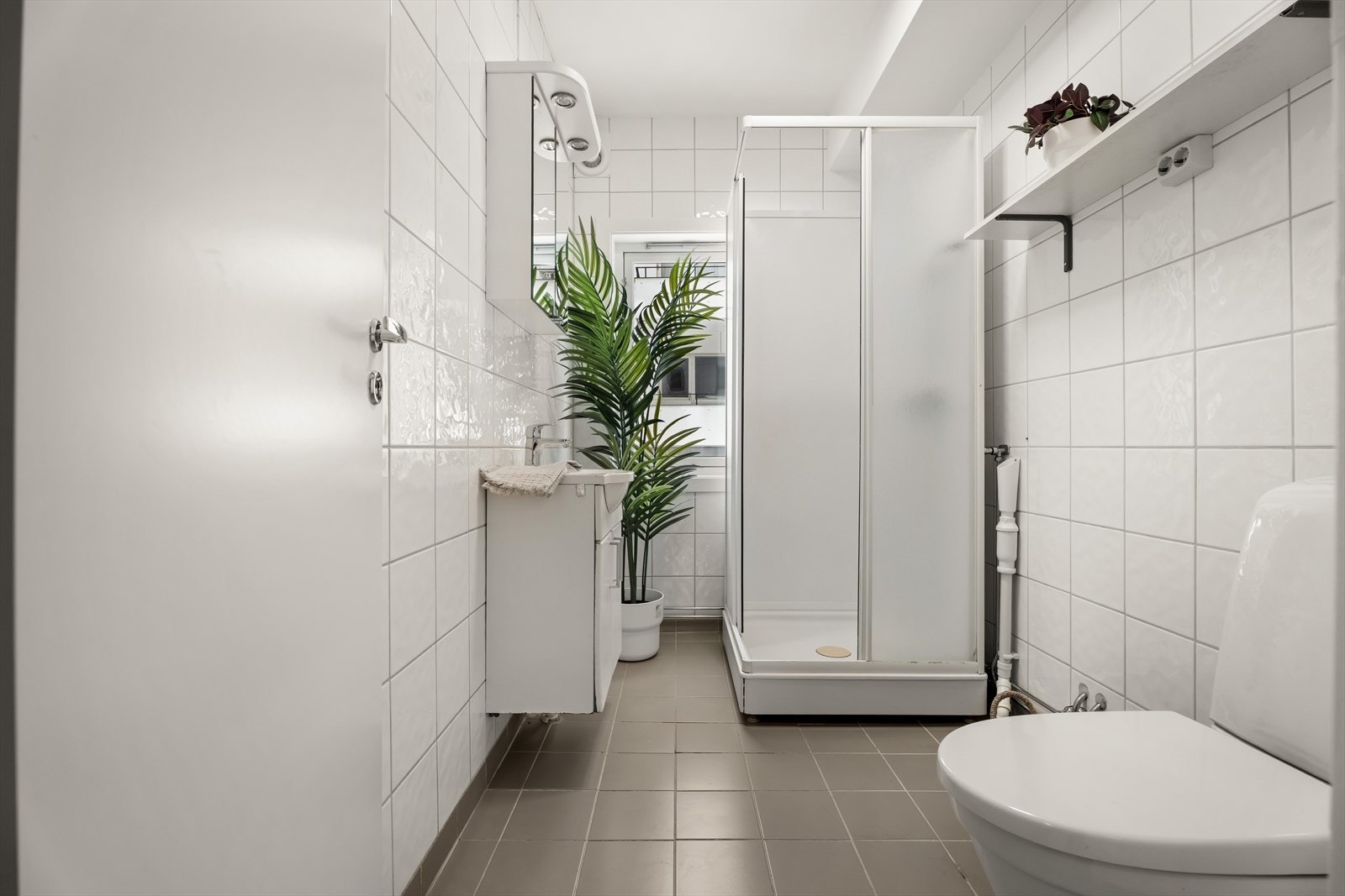 Badet er med fliser på vegger og gulv og malt betong i tak. Videre er det er utstyrt med servantskap, dusjkabinett, opplegg for vaskemaskin og WC på sokkel. Galleribilde