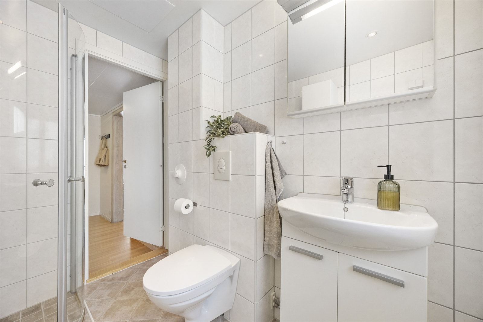 Badet er utstyrt med speilskap med overlys og stikkontakt over servant, dusjhjørne med glassdører og veggmontert dusjarmatur samt vegghengt toalett med innebygget sisterne. Galleribilde