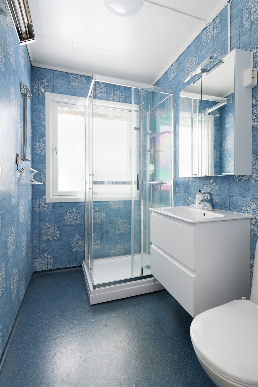 Badet på hovedplan er av eldre dato og har belegg på gulvet og baderomstapet på veggoverflatene. Her er det innredning med dype skuffer og heldekkende servant, samt vegghengt speilskap med overlys. Badet er også innredet med dusjkabinett og toalett. Galleribilde