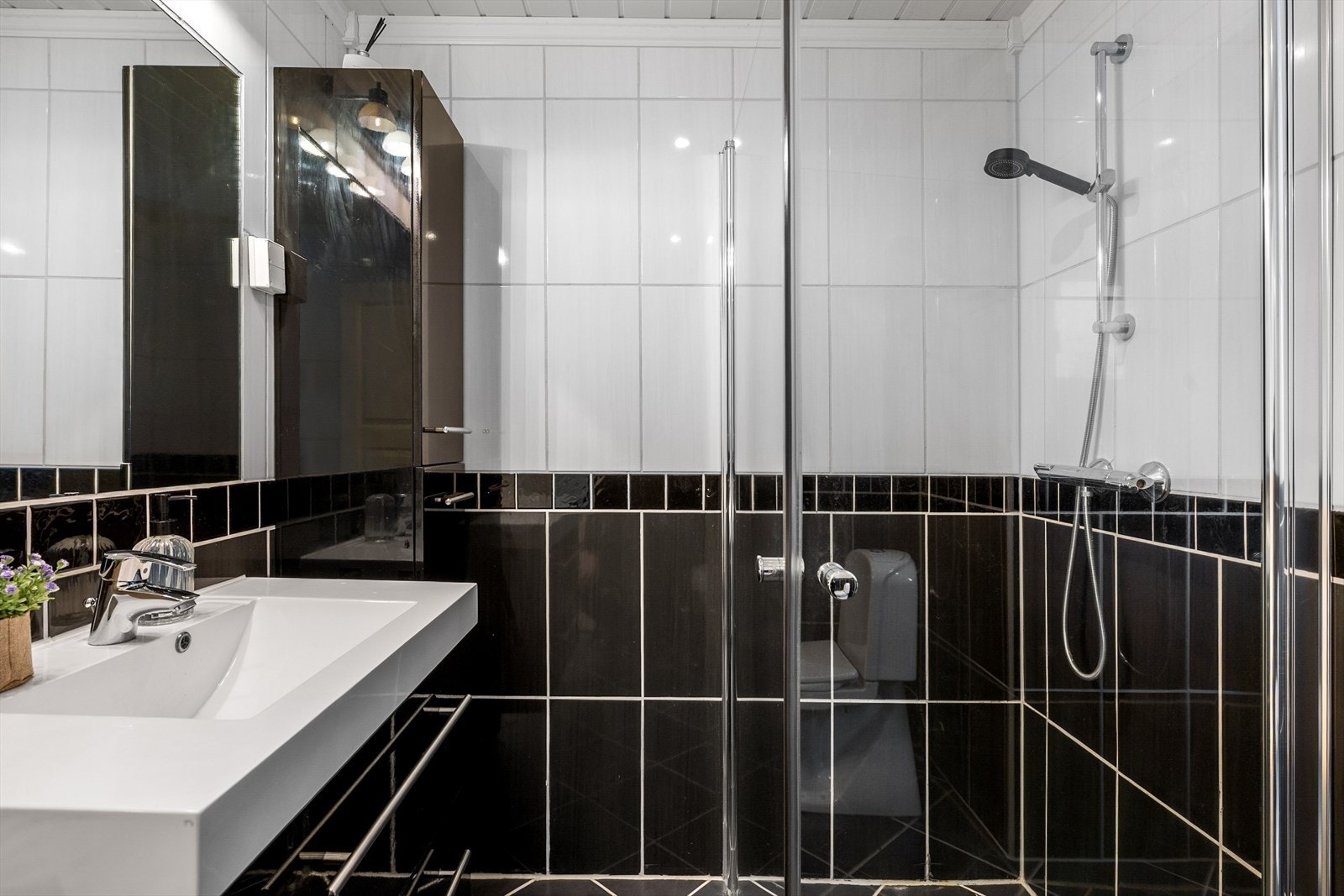 Badet har sort høyglans innredning under heldekkende servant, høyskap og dusjsone bak innfellbare dører. Galleribilde